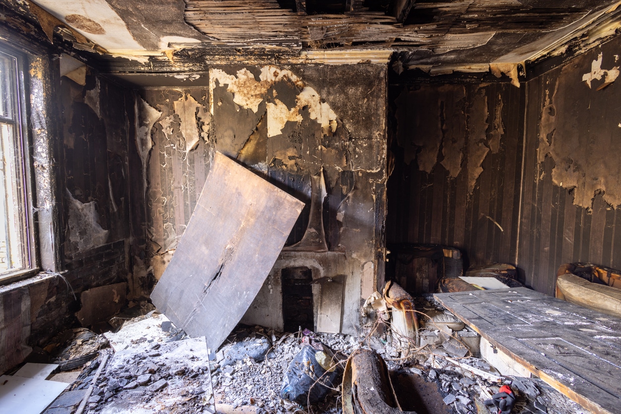 Severely fire-damaged room with charred walls, collapsed ceiling, debris, and a broken fireplace.