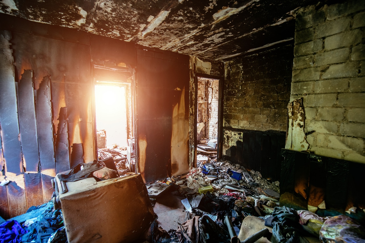Burned-out room with charred walls, debris-strewn floor, and sunlight streaming through a damaged window and doorway.